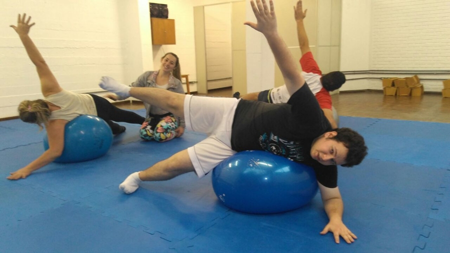 Imagem: Três usuários juntamente com a profissional de educação física, encontram-se em uma sala apropriada para ginástica, onde os usuários estão deitados de lado se equilibrando em cima de uma grande bola, esticando um braço e uma perna em direção ao teto.