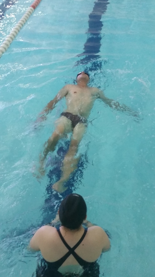 Imagem: Usuário na piscina posicionado com as costas na água e barriga para cima, treinando o nado de costas, sob olhar atento da profissional de educação física. 