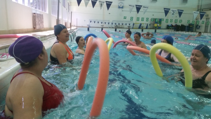Imagem: Grupo misto de usuários dentro da piscina, cada um com uma boia em formato de bastão, segurando cada ponta com uma mão, durante a aula de hidroginástica.