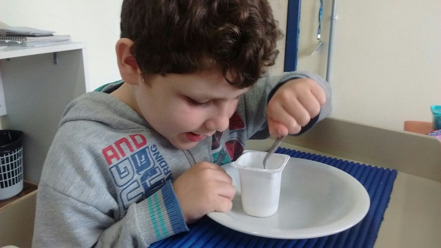 Imagem: Criança sentada em frente à mesa. Sob a mesa encontra-se um prato branco e em cima dela um pote de iogurte aberto. Segurando em sua mão esquerda uma colher, recebendo estimulação para realizar movimentos necessários para levar o alimento à boca. 